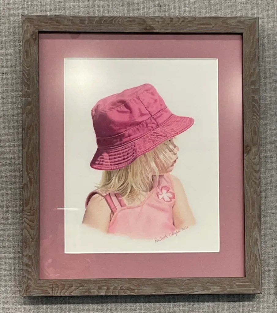 Framed and matted realistic drawing of a two year old girl looking to the side, wearing a pink bucket hat and pink shirt. By Alberta artist Rochelle Colgan