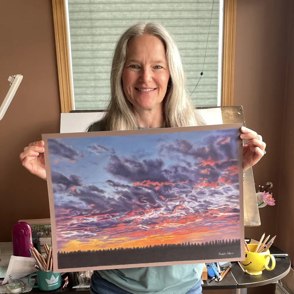 Alberta coloured pencil artist Rochelle Colgan holding her sunset skyscape drawing with purple, pink and orange clouds, light orange sky along the horizon with distant trees