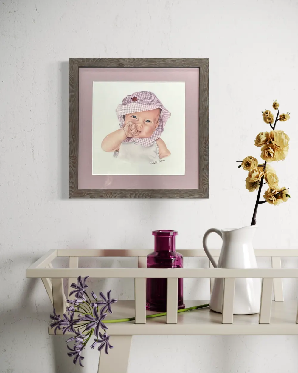 Framed realistic custom portrait of a three month old baby, wearing a gingham pink and white bucket hat and white shirt, hanging on a wall. By Alberta artist Rochelle Colgan