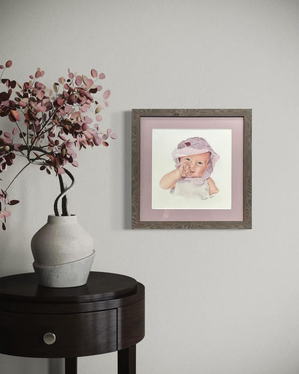 Framed realistic custom portrait of a three month old baby, wearing a gingham pink and white bucket hat and white shirt, hanging on a wall. By Alberta artist Rochelle Colgan