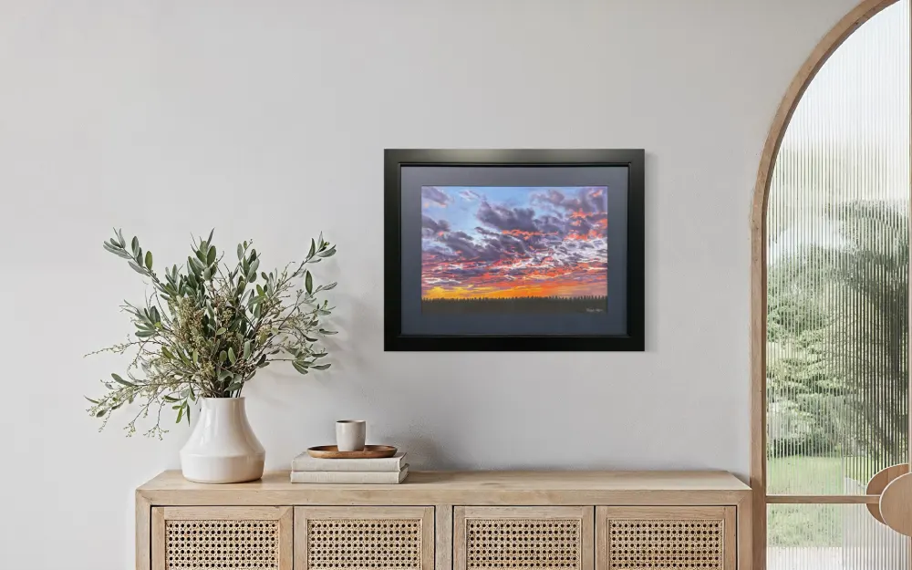 Framed realistic sunset skyscape drawing with purple, pink and orange clouds, light orange sky along the horizon with distant trees, hanging on a wall. By Alberta artist Rochelle Colgan