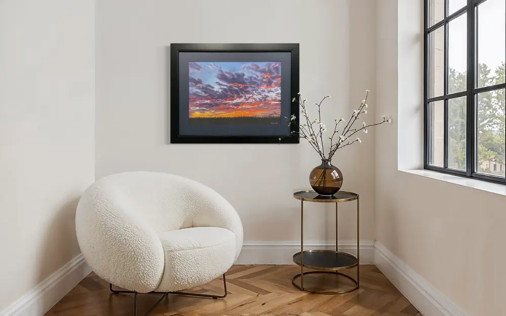 Framed realistic sunset skyscape drawing with purple, pink and orange clouds, light orange sky along the horizon with distant trees, hanging on a wall. By Alberta artist Rochelle Colgan