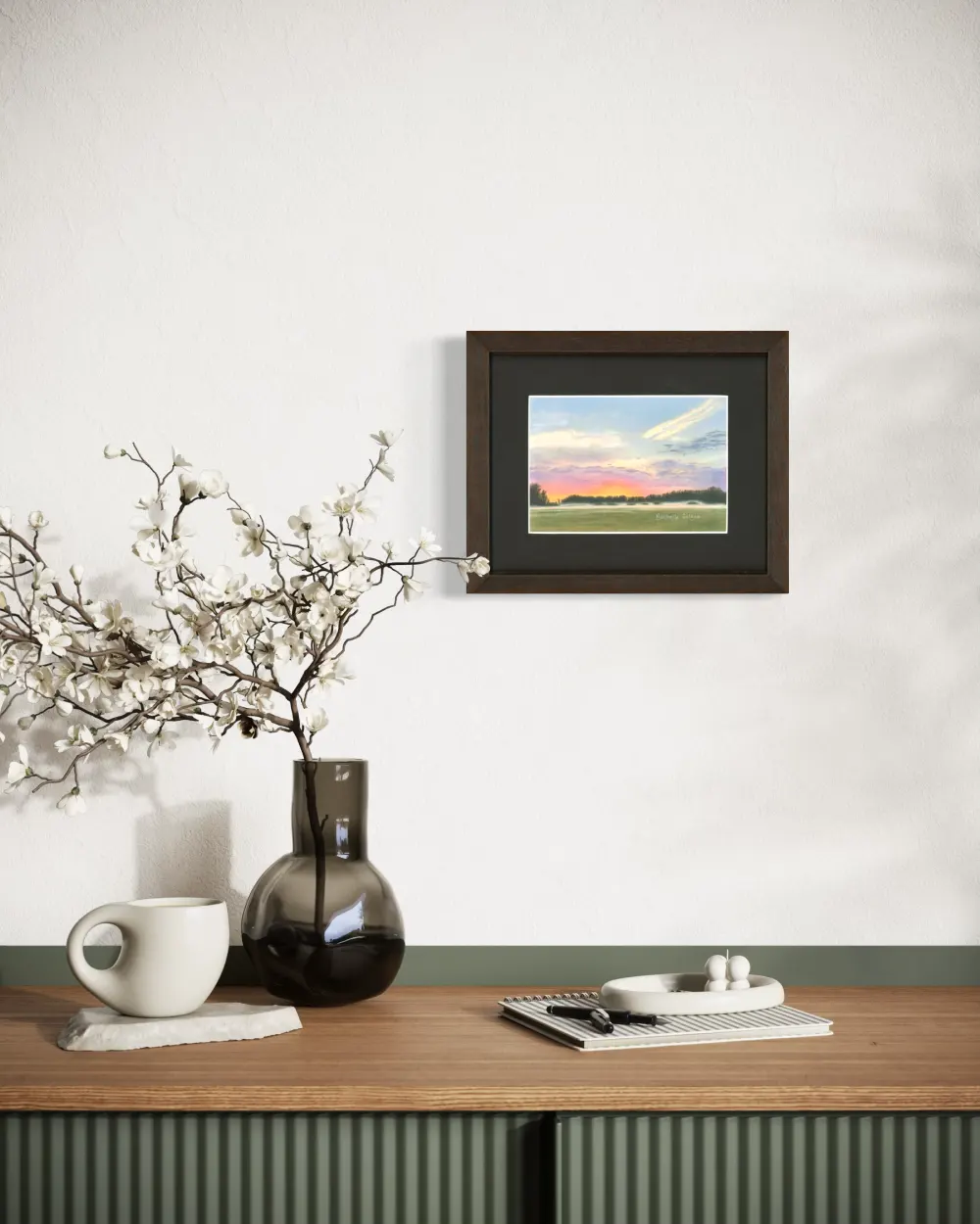 Framed colourful sunrise skyscape drawing with mist, trees and a green grass field, hanging on a wall. By Alberta artist Rochelle Colgan