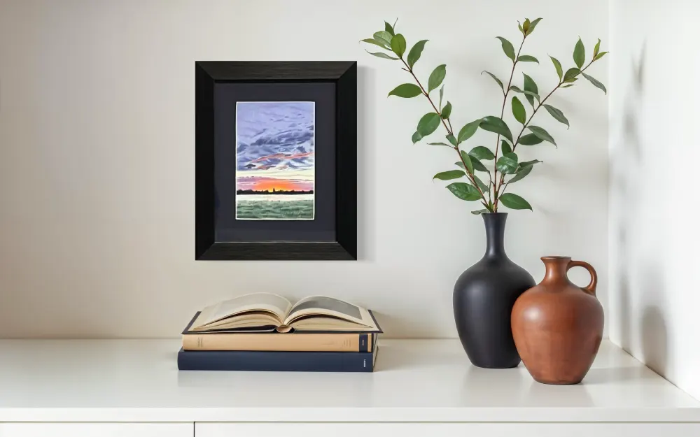 Framed realistic colourful sunrise skyscape drawing with dark clouds, purple and orange horizon, dark trees and mist over a lush green grass field, hanging on a wall. By Alberta artist Rochelle Colgan