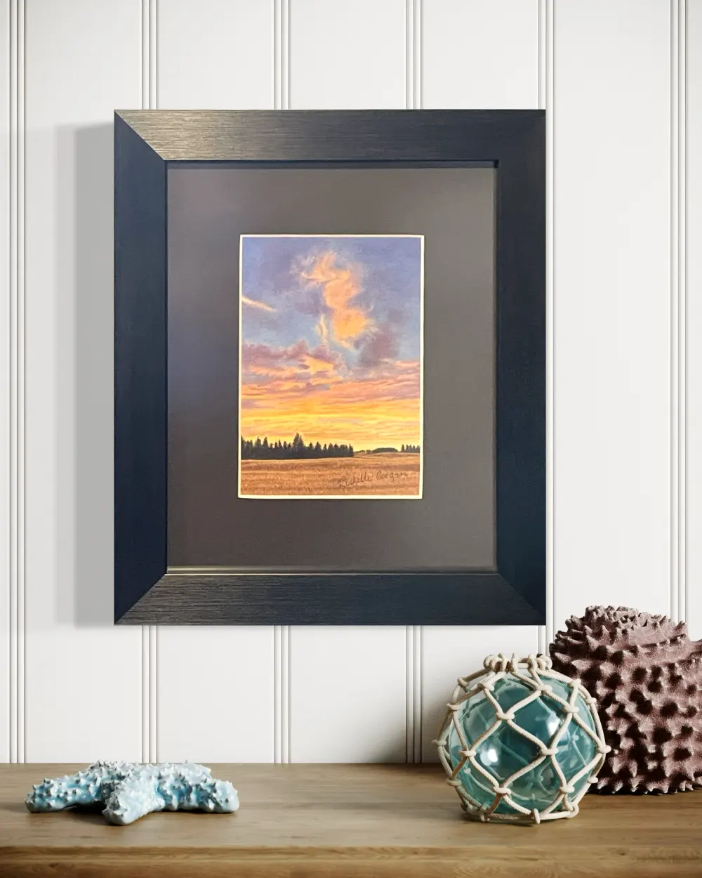 Framed realistic sunrise skyscape drawing of autumn crop field with trees along the horizon, hanging on a wall. By Alberta artist Rochelle Colgan