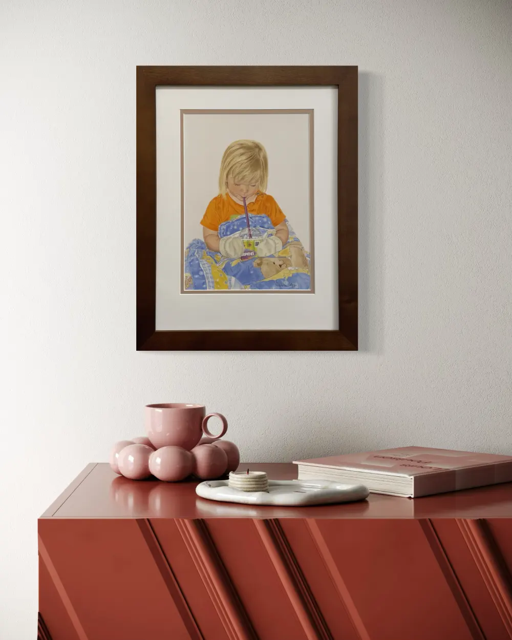 Framed realistic drawing of a three year old girl drinking a slurpee while wearing mittens and covered up with a blanket, hanging on a wall. By Alberta artist Rochelle Colgan