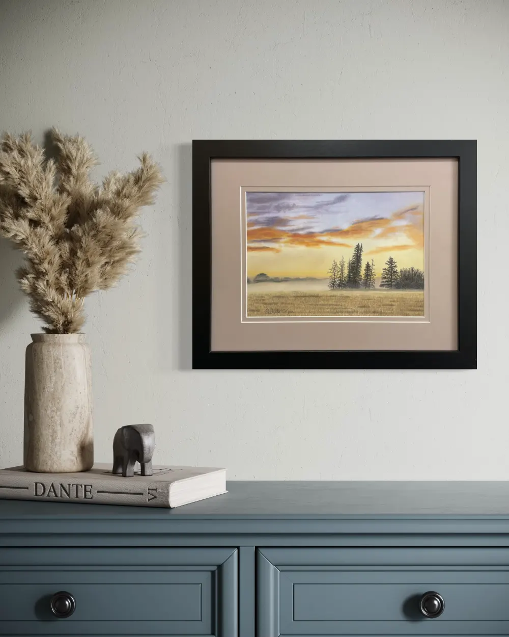 Framed realistic sunrise skyscape drawing with purple and orange clouds, light orange sky along the horizon with distant trees and mist, hanging on a wall. By Alberta artist Rochelle Colgan